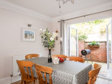 A dining room with wooden chairs around a table covered with a grey tablecloth and a vase of flowers at Mews Cottage in Menai Bridge