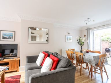 A living room with a grey sofa red and white cushions a dining table with chairs and a vase with flowers at Mews Cottage in Menai Bridge
