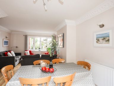 A dining area with a table covered by a cloth and chairs and a living room with sofas and armchairs near a window at Mews Cottage Menai Bridge