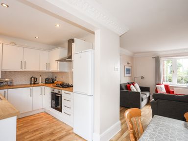 A kitchen with white cabinets and wooden countertops next to a living room with two gray sofas and a red armchair at Mews Cottage in Menai Bridge