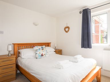 A bedroom with a wooden bed white bedding two rolled towels on the bed two wooden nightstands with lamps and a window with dark curtains at Mews Cottage in Menai Bridge