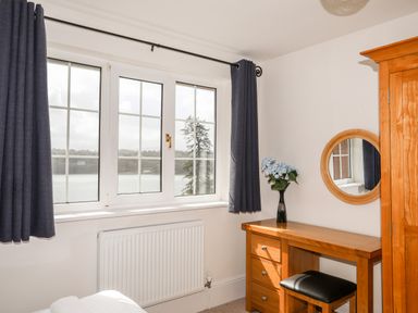 A room with a window with curtains a wooden dressing table with a mirror and a vase of flowers and a wooden wardrobe at Mews Cottage in Menai Bridge