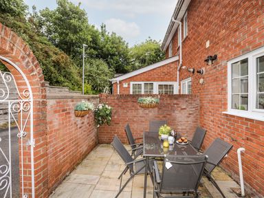 An outdoor patio area with a glass dining table, six chairs, and hanging flower baskets at Mews Cottage in Menai Bridge