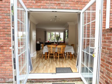 An open glass door leading to a dining area with a table covered by a patterned cloth surrounded by wooden chairs at Mews Cottage in Menai Bridge