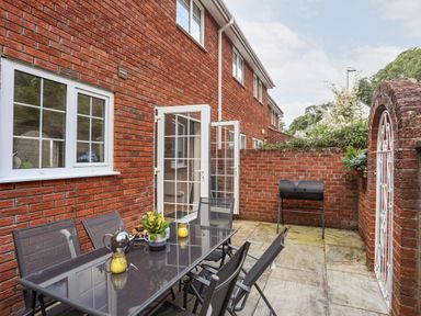 An outdoor patio area with a glass table and six chairs a barbecue grill and open double doors at Mews Cottage in Menai Bridge