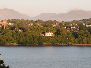A view of a river with trees and houses on the opposite bank and mountains in the background at Mews Cottage in Menai Bridge