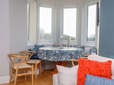 A dining area with a round table covered with a blue patterned tablecloth surrounded by wooden chairs and cushions at Min Y Mor Trearddur Bay