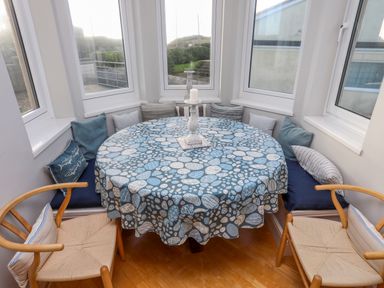 A dining area with a round table covered with a patterned tablecloth surrounded by built-in cushioned seating and wooden chairs at Min Y Mor Trearddur Bay