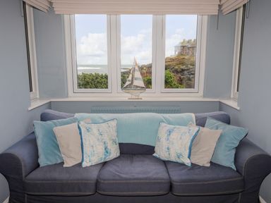 A blue sofa with multiple cushions and a small sailboat decoration on the windowsill with an ocean view at Min Y Mor Trearddur Bay