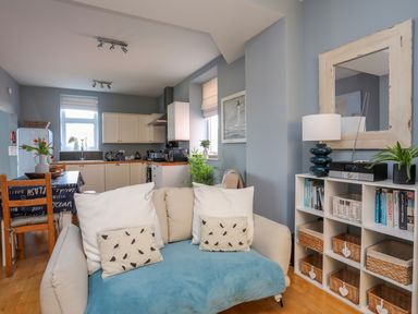 A kitchen and living area with a beige sofa, dining table with chairs, white cabinets, and a shelving unit with books and baskets at Min Y Mor Trearddur Bay in Trearddur Bay