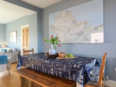 A dining area with a wooden table covered with a navy blue tablecloth with white text a vase with flowers bread and grapes on the table a large map on the wall and part of a living room with striped furniture in the background at Min Y Mor Trearddur Bay