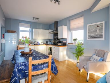 A kitchen and dining area with a table covered by a blue tablecloth with chairs a vase of flowers a light blue refrigerator white cabinets cooking appliances and an armchair with a cushion and a plant near a window at Min Y Mor Trearddur Bay in Trearddur Bay