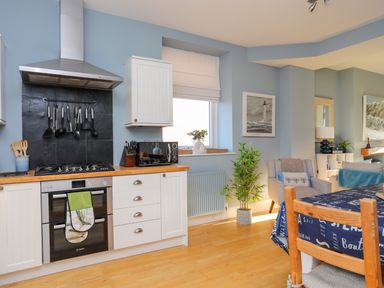 A kitchen with white cabinets a stove and oven a dining table with a blue cloth and a gray armchair at Min Y Mor Trearddur Bay in Trearddur Bay