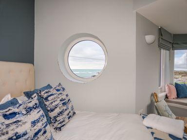 A bedroom with a round window showing the sea and pillows on a bed at Min Y Mor Trearddur Bay
