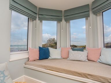 A bay window seating area with cushions overlooking the sea and a house on a rocky hill at Min Y Mor Trearddur Bay in Trearddur Bay