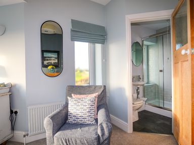 A room with a gray armchair and two cushions near a window and a bathroom with a shower at Min Y Mor Trearddur Bay