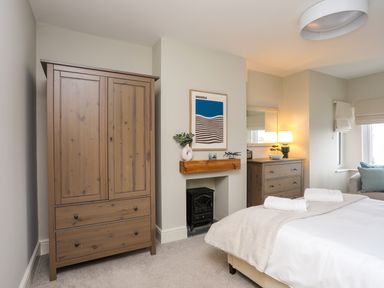 A bedroom with a wooden wardrobe, a fireplace with a shelf and decorations, a chest of drawers with a lamp, and a bed with towels at Min Y Mor Trearddur Bay