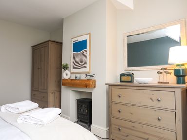A bedroom with a wooden wardrobe a wooden chest of drawers with a mirror and lamp and a small black stove in a recess in the wall at Min Y Mor Trearddur Bay