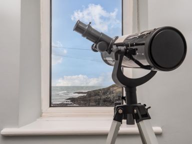 A telescope on a tripod by a window with a view of the sea and rocky coastline at Min Y Mor Trearddur Bay in Trearddur Bay