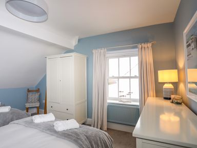 A bedroom with two beds a white wardrobe a chair under a slanted ceiling a window with white curtains a dresser with a lamp and decorations at Min Y Mor Trearddur Bay in Trearddur Bay