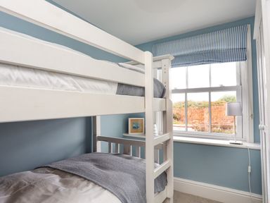 A bedroom with white bunk beds a window with striped curtains and a lamp on the windowsill at Min Y Mor Trearddur Bay