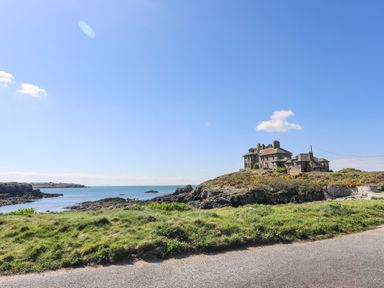 A stone house on a rocky hill by the sea with a grassy area and a road in the foreground at Min Y Mor Trearddur Bay