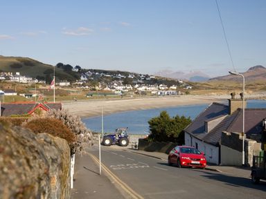 A coastal road with a red car and a tractor near a beach with houses on the hillside at Morfin in Criccieth