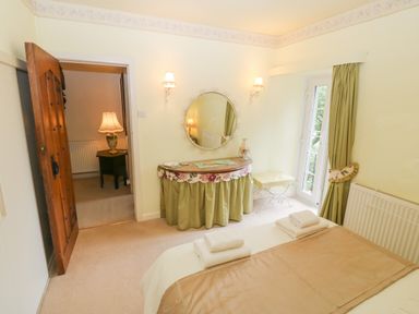 A bedroom with a bed with folded towels a wooden door a round mirror above a draped table and a bench by a window with curtains at Morfa Lodge in Porthmadog