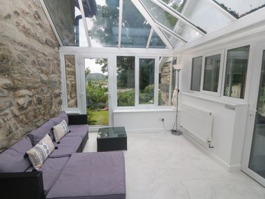 A conservatory with a glass ceiling stone wall grey sofa with cushions glass coffee table white floor lamp and garden view at Morfa Lodge in Porthmadog