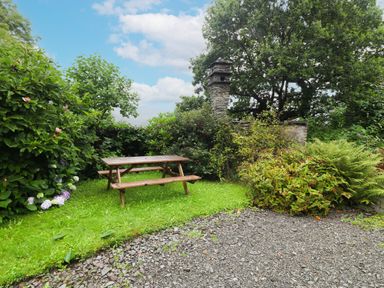 A wooden picnic table on grass surrounded by bushes and trees with a stone chimney in a garden