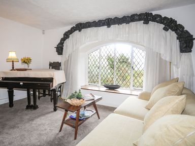 A living room with a piano table with lamp flowers and sofa near a window with white curtains at Morfa Lodge in Porthmadog
