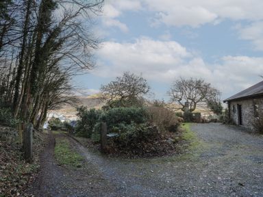 A gravel driveway next to a stone building with trees and bushes around and hills in the background at Morfa Lodge in Porthmadog