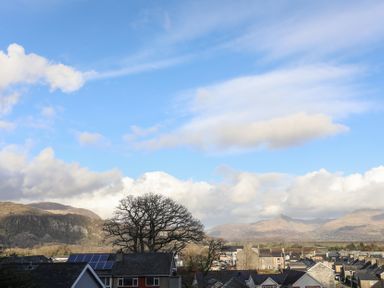 A residential area with rooftops and a large tree in front of mountains under a blue sky with clouds at Morfa Lodge in Porthmadog