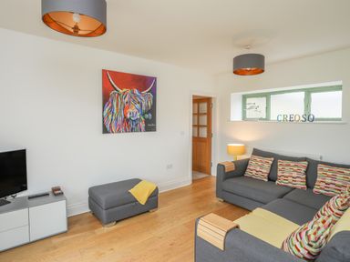 A living room with a gray sectional sofa and colorful cushions a gray ottoman wooden flooring and a colorful cow painting on the wall at Mynydd Parys in Brynsiencyn