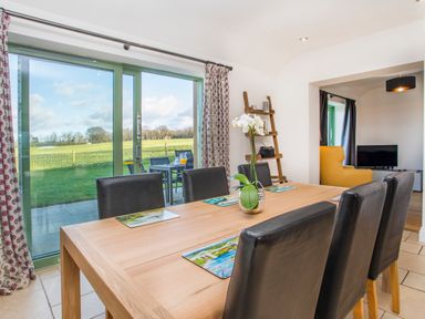 A dining area with a wooden table and black chairs near sliding glass doors overlooking a green field at Mynydd Parys in Brynsiencyn
