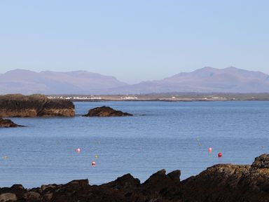 A coastal scene with water, rocks, buoys, distant houses, and mountains at Pant yr Hyman in Rhoscolyn