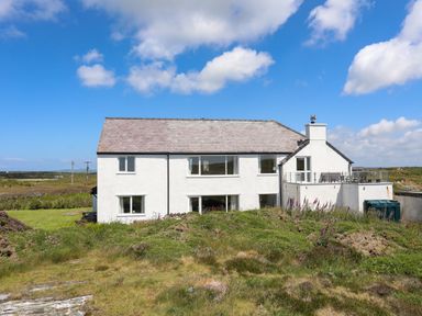 A white two-story house with a slate roof and glass balcony surrounded by grass and shrubs at Pant yr Hyman in Rhoscolyn