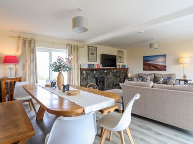 A dining area with a wooden table and white chairs next to a living room with beige sofas a stone fireplace and a television at Pant yr Hyman in Rhoscolyn