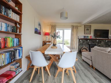 A dining area with a wooden table and white chairs next to a bookshelf and a living room with a stone fireplace at Pant yr Hyman in Rhoscolyn