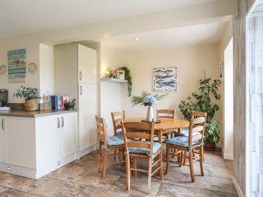 A dining area with a round wooden table and six chairs with blue cushions next to kitchen cabinets and plants at Pant yr Hyman in Rhoscolyn