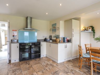 A kitchen with a black stove and oven a dining table with wooden chairs and blue cushions at Pant yr Hyman in Rhoscolyn