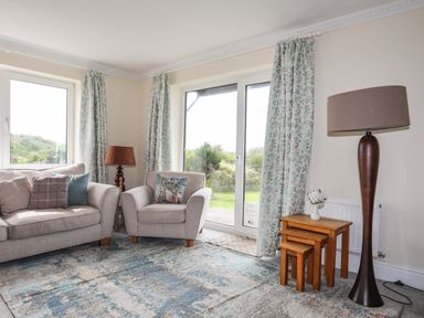 A living room with sofa armchair floor lamp nested wooden tables and large windows with curtains at Pant yr Hyman in Rhoscolyn