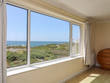 A room with large windows overlooking greenery and the sea with curtains and a wooden chest at Pant yr Hyman in Rhoscolyn