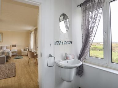 A bedroom with a bed and chair next to a bathroom sink and window with curtain at Pant yr Hyman in Rhoscolyn