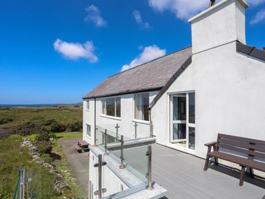 A balcony with a wooden bench and glass railing on a white house with a view of greenery and the sea at Pant yr Hyman in Rhoscolyn