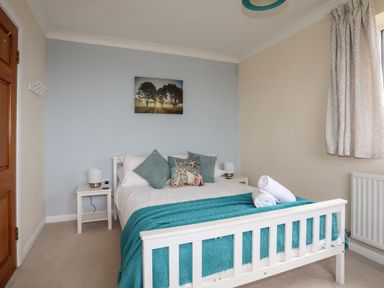 A bedroom with a white bed with teal blanket and pillows two bedside tables with lamps a wooden door and a window with curtains at Pant yr Hyman in Rhoscolyn