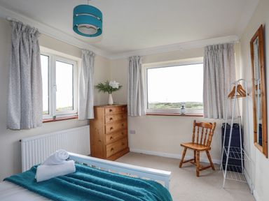 A bedroom with two windows wooden chest of drawers wooden chair clothes rack and bed with teal blanket at Pant yr Hyman in Rhoscolyn