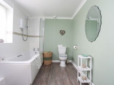 A bathroom with a bathtub shower combo a toilet a wicker laundry basket a wall mirror and a small white shelf at Pant yr Hyman in Rhoscolyn