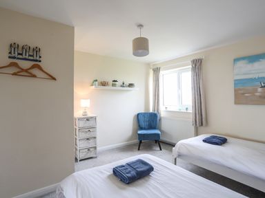 A bedroom with two single beds each with a folded towel on top a blue chair by the window a small chest of drawers with a lamp on it and a shelf with small plants and decor at Pant yr Hyman in Rhoscolyn