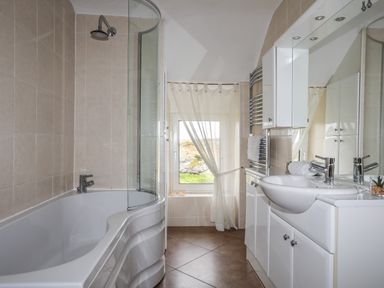 A bathroom with a bathtub shower combination a window with sheer curtains and a sink with cabinets at Pant yr Hyman in Rhoscolyn